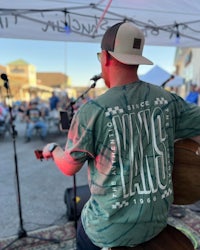 a man in a t - shirt sitting on a stool at an outdoor event